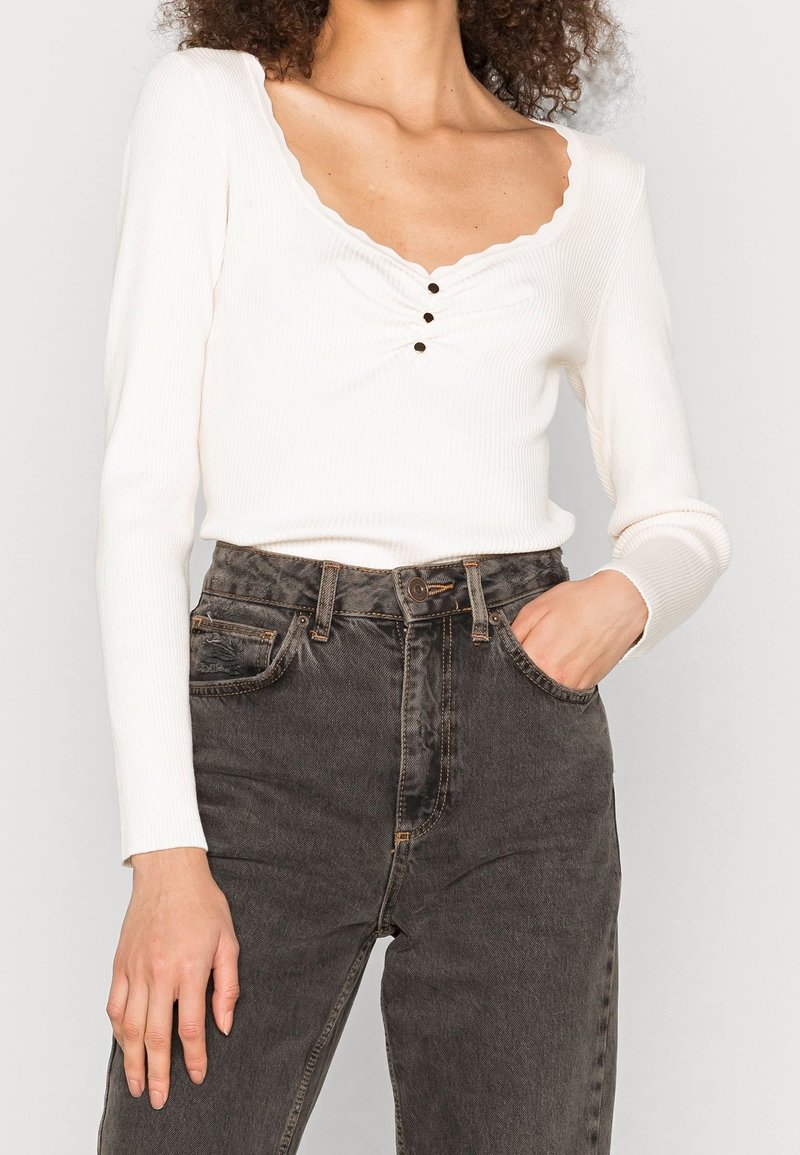 Maglietta bianca a maniche lunghe con colletto ondulato e tre bottoni neri decorativi, abbinata a jeans grigi scuri a vita alta.