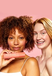 Kaksi naista hymyilee vaaleanpunaista taustaa vasten. Toinen pitää kädessään sylinterimäistä persikanväristä putkea, jossa lukee "INDIE LEE". Molemmilla on luonnollinen, raikas ilme.