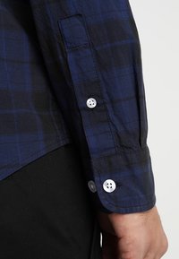 Camicia a maniche lunghe blu e nera a scacchi con bottoni bianchi ai polsini; materiale in cotone; presenta una vestibilità comoda e dettagli di cucitura visibili.