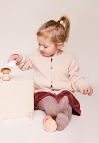 Houten speelgoedpaddenstoelen in natuurlijke en bruine tinten, met een kind dat ermee speelt. Het kind draagt een beige vest en een bordeauxrode jurk, zittend op een lichte achtergrond.