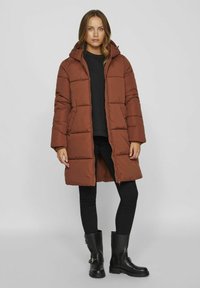 Doudoune marron avec une capuche, présentant des coutures horizontales, des poches avant et une fermeture éclair, associée à un jean skinny noir et des bottes.