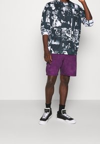 Schwarz-weißes grafisches Langarmshirt, lila Batikshorts und schwarze High-Top-Sneaker mit weißen Schnürsenkeln und Akzenten.