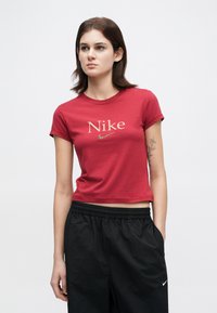 Νεαρή γυναίκα φορώντας ένα κόκκινο κοντό μπλουζάκι Nike και μαύρο παντελόνι Nike, στέκεται με το ένα χέρι στην τσέπη και ένα λουλουδάτο τατού στο χέρι της.