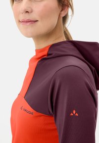 Roter und lila Fleece-Hoodie mit hohem Kragen, der ein Logo auf der Brust und ein Emblem am linken Ärmel trägt. Glatte Textur, verstellbare Kapuze.