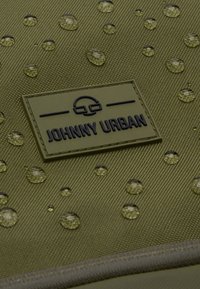 Olivově zelená tkanina s texturou, na které jsou kapky vody a vyvýšená černá nášivka s nápisem "JOHNNY URBAN."