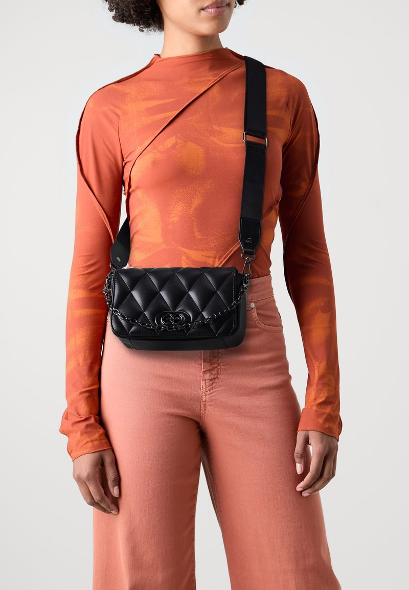 Femme portant un haut orange à manches longues et un pantalon rose, portant un sac bandoulière noir matelassé avec chaîne et large sangle.