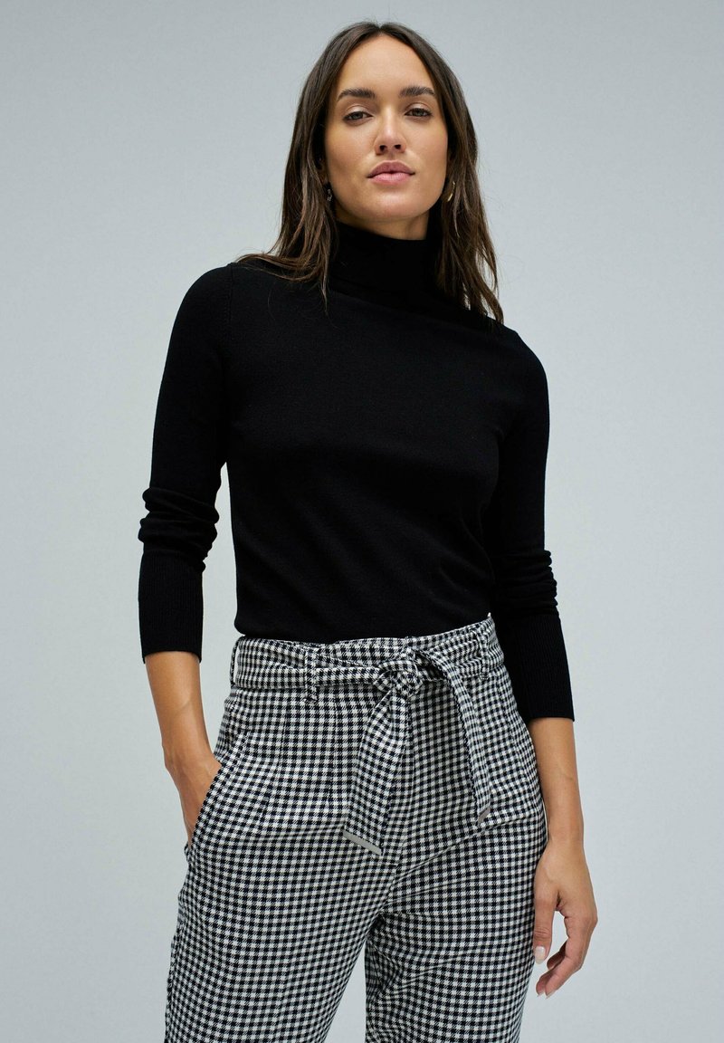Pull à col roulé noir à manches longues associé à un pantalon taille haute à carreaux noir et blanc avec une ceinture nouée.