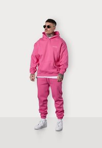 Un sweat à capuche rose vif et un jogging assorti fabriqués en tissu doux. Le sweat à capuche a une poche avant et des poignets en côte. Porté avec des baskets blanches.