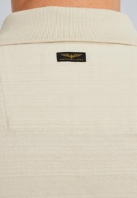 Lys beige polo skjorte med ribbet krave, tekstureret stof og en sort etiket med guld skrift på ryggen.
