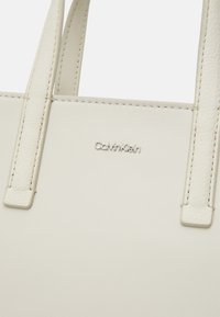 Bela usnjena torbica z dvema zgornjima ročajema. Vključuje embosirano oznako 'Calvin Klein'. Gladka tekstura z minimalističnim dizajnom in ojačanimi šivi.