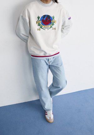 Osoba oblečená v bílo-šedé mikině Adidas s barevným logem, světle modrých džínách a bílých teniskách s červenými pruhy, stojící na modré podlaze.