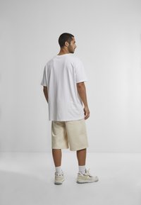 White cotton t-shirt, relaxed fit, paired with light beige shorts. Sneakers with white and yellow accents, plain background.