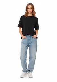 T-shirt nero a maniche corte abbinato a jeans larghi di colore blu chiaro. Il modello sta in piedi con le mani in tasca, indossando sneakers bianche.