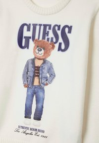Sweat-shirt blanc avec un graphisme d'ours vêtu en denim, avec le mot « GUESS » en lettres majuscules, et un texte de marque en bas.