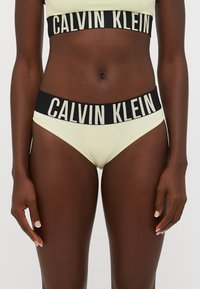 Calvin Klein gele bralette en bijpassende slips met zwarte elastische banden die voorzien zijn van witte merknaamletters. Glad materiaal, klassieke snit.