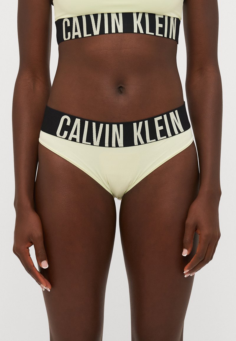 Calvin Klein gele bralette en bijpassende slips met zwarte elastische banden die voorzien zijn van witte merknaamletters. Glad materiaal, klassieke snit.
