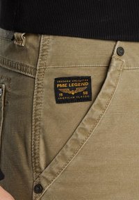 Beige cargo pants with a black and yellow PME Legend label, featuring stitched detailing, front pockets, and a textured fabric finish.