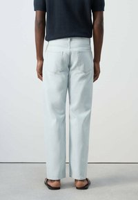 Persona con vaqueros azul claro de corte recto, camiseta negra de manga corta y sandalias negras, de espaldas sobre un fondo liso.