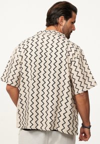 Kurzarmhemd aus cremefarbenem Stoff mit schwarzen Zickzack-Mustern, Kragen und lockerer Passform. Sichtbares strukturiertes Webdetail.