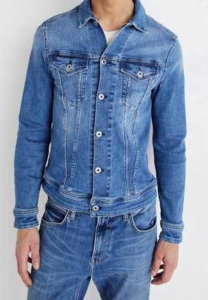 Person trägt eine blaue Jeansjacke mit Knöpfen und Fronttaschen sowie passende blaue Jeans, steht vor weißem Hintergrund.