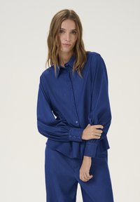 Camicia blu con bottoni, maniche lunghe a pieghe e una vestibilità rilassata. Realizzata in tessuto liscio con un leggero lucido, con dettagli di bottoni.
