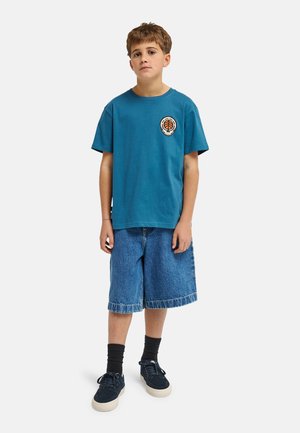 Bawełniany turkusowy t-shirt z okrągłym logo, zestawiony z niebieskimi szortami dżinsowymi o długości do kolan z postrzępionym wykończeniem. Obuwie to ciemne trampki.