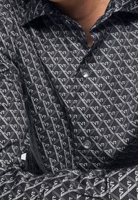 Černá košile s geometrickým vzorem bílých trojúhelníků a písmen, s knoflíkovým zapínáním a texturovanou tkaninou.
