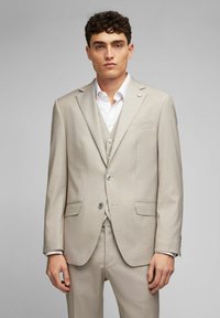 Helles beige maßgeschneidertes Anzug mit einem einreihigen Sakko, zwei vorderen Taschen, einem Reverskragen und einer subtilen Textur. Darunter ein weißes Hemd.