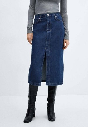 Femme portant une jupe midi en denim bleu foncé avec une fente à l'avant, un haut gris à manches longues et des bottes noires à talons montant jusqu'aux genoux, debout sur un sol blanc.