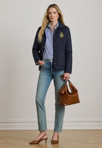 Μπλε ναυτικό ρουμπουλό μπουφάν με χρυσό έμβλημα, συνδυασμένο με μπλε και άσπρο ριγέ πουκάμισο, ανοιχτόχρωμο τζιν, καφέ δερμάτινη τσάντα και τακούνια.