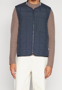 Marinblått vadderad väst med quiltat mönster, dragkedja framtill och bröstficka, bärs över en ljusbrun tröja och krämfärgade byxor.