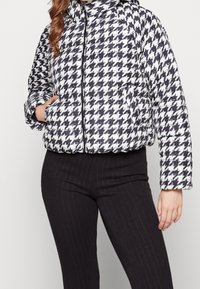 Kvinna iförd en svart och vit houndstooth pufferjacka med dragkedja och svarta ribbad leggings, hand i jackfickan.