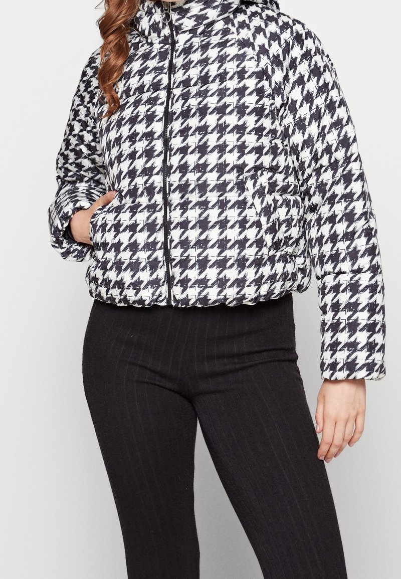 Kvinna iförd en svart och vit houndstooth pufferjacka med dragkedja och svarta ribbad leggings, hand i jackfickan.