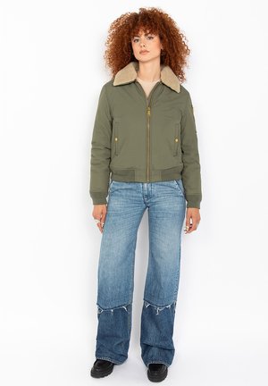 Giubbotto bomber verde oliva con colletto in pelliccia sintetica beige, chiusura a zip e orlo a coste. Abbinato a jeans a gamba larga blu chiaro con bordi sfrangiati.