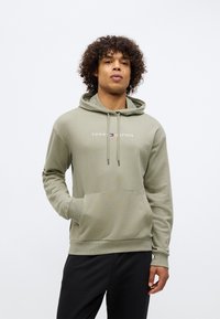 Mladý muž s kučeravými vlasmi, oblečený v svetlozelenej mikine Tommy Hilfiger a čiernych nohaviciach, stojí s jednou rukou v vrecku.