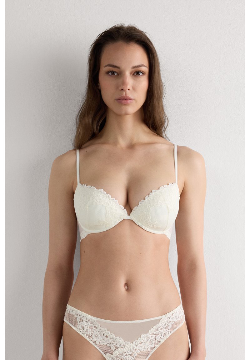 Woman with long brown hair wearing matching white lace bra and panties standing against a plain light wall, facing forward.
