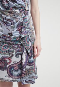 Ärmlös klänning med ett ljusblått tyg som har intrikata lila och gröna paisleymönster. Inkluderar en dekorativ bältesspänne vid midjan.