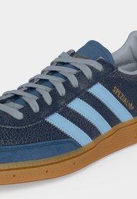 Αθλητικό παπούτσι Adidas Spezial σε ναυτικό μπλε με ανοιχτό μπλε ρίγες, γκρι κορδόνια, υφασμένο επάνω μέρος και σόλα από καουτσούκ, εμφανιζόμενο σε ελαφριά γωνία.