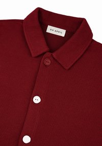 Camicia button-up bordeaux con una texture a coste. Presenta un colletto e due bottoni bianchi, etichettati "OH APRIL" all'interno del colletto.