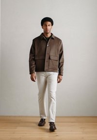 Pruun, nööbitav jakk, mis on sileda tekstuuriga, kahe ees taskuga ja kohandatud lõikega, kantud valgete pükste ja tumedate kingadega.