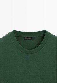 Sweat-shirt vert foncé à col rond côtelé avec une petite étiquette noire cousue à l'intérieur du col.