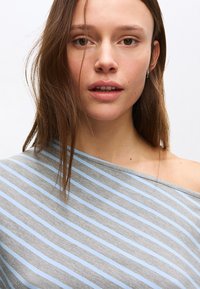 Giovane donna con capelli castano chiaro, che indossa una maglietta a righe grigia e azzurra con spalle scoperte, che guarda dolcemente verso la fotocamera su uno sfondo bianco.