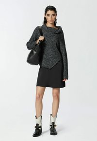 Maglione nero a maglia con collo a pipistrello, abbinato a un vestito di raso nero e stivaletti neri e bianchi. La modella tiene in mano una borsa nera.