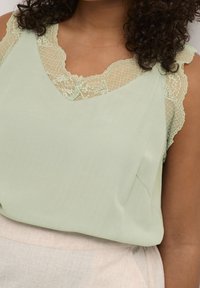 Light green sleeveless top with lace accents at shoulders, featuring a deep V-neck and a soft, textured fabric. Worn with a neutral bottom.