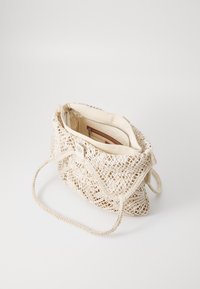 Sac à main beige tissé avec un extérieur texturé, une poignée en corde et une fermeture par bouton. L'intérieur comprend une poche zippée et une doublure en tissu.
