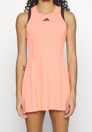 Frau trägt ärmelloses sportliches Kleid in Korallenrosa mit sichtbaren schwarzen Sport-BH-Trägern und kleinem schwarzem Adidas-Logo auf der Brust.