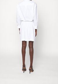 Abito camicia bianco a maniche lunghe con vita aderente, pieghe sul retro e orlo arrotondato, abbinato a tacchi neri con cinturini.