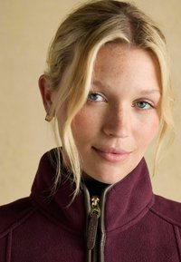 Burgunderfarbene Fleecejacke mit Reißverschluss und hohem Kragen, ausgestattet mit braunen Beschlägen und einer strukturierten Oberflächenbeschaffenheit. Der helle Hintergrund hebt die Farbe des Kleidungsstücks hervor.