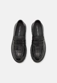 Mocassins en cuir noir avec un motif crocodile texturé, présentant un design classique à enfiler et un logo discret sur la semelle intérieure.