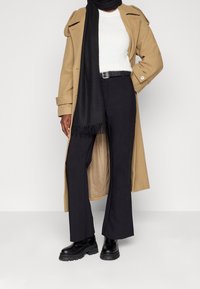 Person i beige lång kappa, svart scarf, vit tröja, svarta byxor med bälte och svarta kraftiga stövlar på en neutral bakgrund.